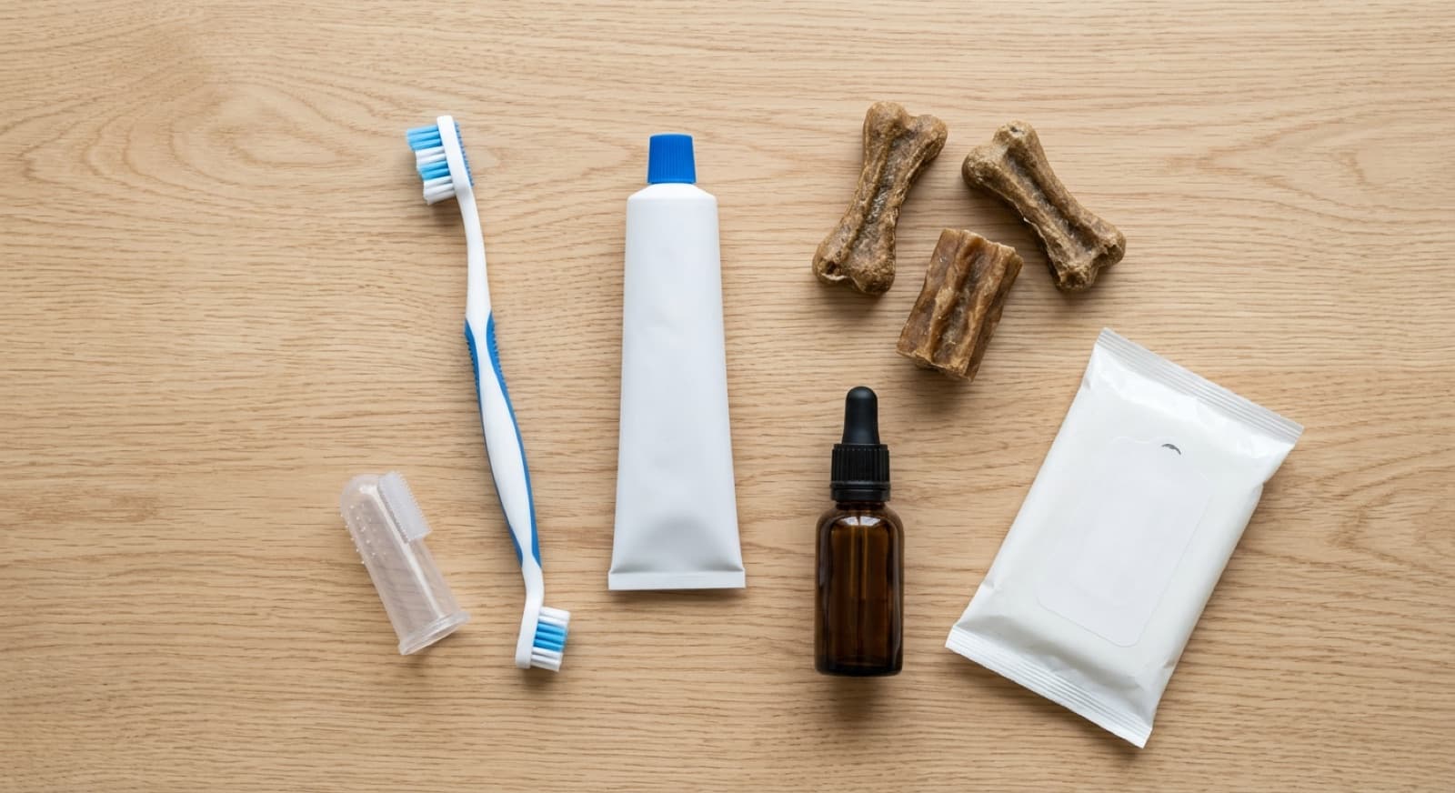 A flat-lay of the home dog-dental toolkit: a soft dog toothbrush and finger-brush, a tube of dog toothpaste, dental chews, a bottle of water additive, and dental wipes